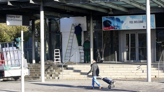 Arbeiter schrauben nach dem Brandanschlag am Eingang der Messehallen Gipsplatten vor eine beschädigte Glasscheibe.