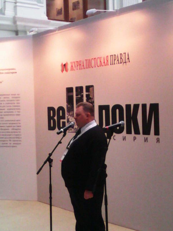 Vladislav Shurigin bei der Eröffnung von "Beweise. Syrien" im Dezember 2013 in Moskau. Im Hintergrund: Schriftzug der ultranationalistischen Žurnalistskaja Pravda (dt. „Journalistische Wahrheit“)