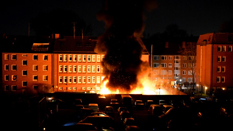 Die Flammen aus dem Hinterhof stehen meterhoch. Autos auf dem angrenzenden Marktplatz sind durch eine Mauer vor den Flammen geschützt.